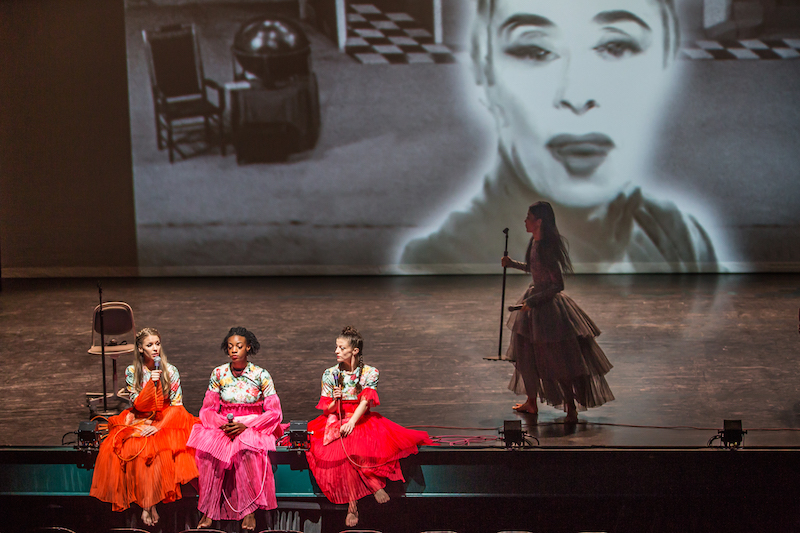 Three women in brightly colored dresses sit on the edge of the stage. One woman talks into a microphone. Xin Ying, in a grey colored dress stands to the right of them holding a microphone stand. A projection of Martha Graham is in the background.